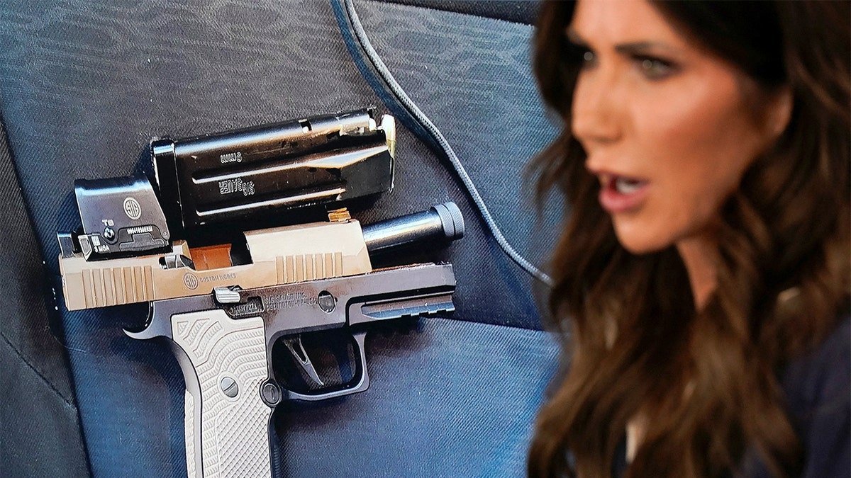 A cabinet official addresses reporters as a firearm image is displayed behind her on a large screen.