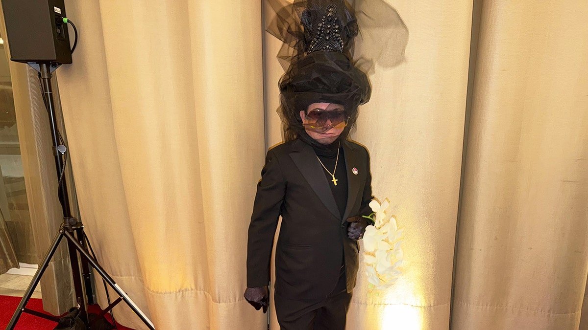 Andre Soriano Atelier dressed in an all-black formal suit with an oversized sculptural headpiece, holding white flowers at "Melania" premiere.