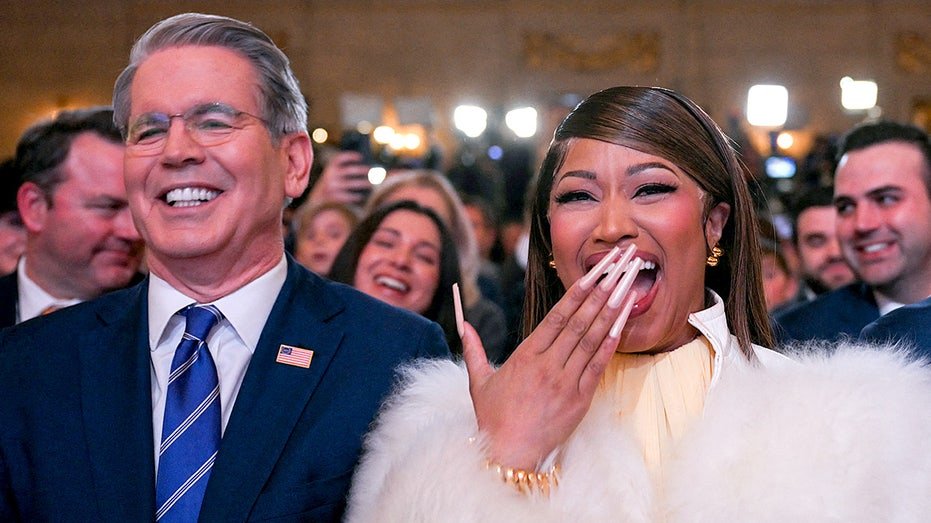 Scott Bessent and Nicki Minaj stand listening as Donald Trump speaks at a podium during an event.